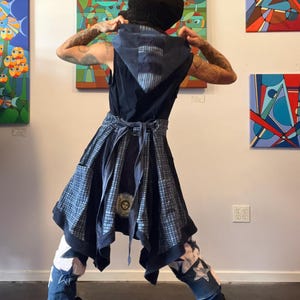 May include: A person wearing a black hooded vest and a patterned skirt with a tie-waist. The outfit includes star-patched jeans and black boots. Colorful abstract paintings hang on the wall in the background.