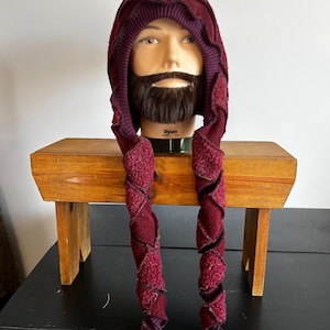 May include: A burgundy hooded hat with long, spiraling tassels. The hat is made of a textured fabric and features a ribbed knit trim. The hat is displayed on a mannequin head with a beard. The word "Dylan" is printed on the base of the mannequin.