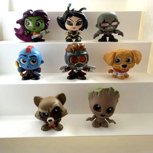 May include: Nine character figurines from a science fiction franchise are displayed on white shelves. The figures include a raccoon, a dog, and a tree-like creature, each with unique designs and colours. The figurines are approximately 7-10 cm tall.