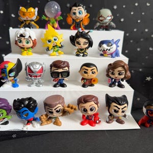 Puede incluir: Una colección de coloridas figuras de superhéroes exhibidas en una plataforma blanca escalonada. Las figuras presentan varios personajes con disfraces y expresiones distintas. El fondo es negro con acentos en forma de estrella blanca.