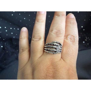 May include: A silver-colored ring in the shape of a skeleton hand. The ring is worn on a finger and features detailed bone structures. The ring is a gothic or alternative style accessory.