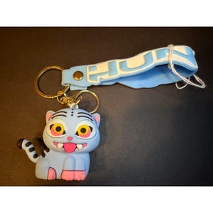 May include: A light blue silicone keyring featuring a cartoon tiger character with yellow eyes, a pink tongue, and white fangs. Attached to the gold-toned metal ring is a light blue silicone strap with the white text HUN printed on the surface.
