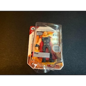May include: A Thor action figure encased in clear plastic packaging on a cardboard backing. The figure features blonde hair, a grey and black suit, and a red cape. The packaging includes the text Thor and a circular label stating hammer included.