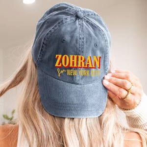 May include: Navy blue baseball cap with the text "ZOHRAN for NEW YORK CITY" in yellow and red lettering. The cap is made of a soft, worn material.