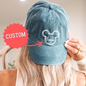 May include: Teal baseball cap featuring a white Mickey Mouse outline and the name "Sarah". The cap has a curved peak and a worn appearance. The word "CUSTOM" is in a red starburst shape.