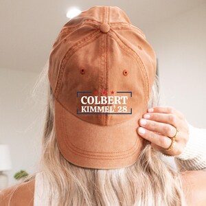 May include: A Texas Orange baseball cap with the text "COLBERT KIMMEL 28" in a blue and red design. The cap is a solid color and is being worn by a person with long blonde hair.