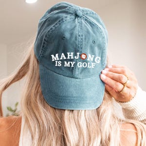 May include: Teal baseball cap with the text "MAHJONG IS MY GOLF" in white lettering. The cap has a curved brim and a classic six-panel design. The cap is made of a soft, washed cotton material.