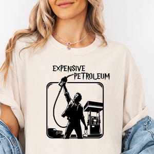 May include: A white t-shirt with a black graphic of a person pumping gas at a gas station. The graphic says "Expensive Petroleum".