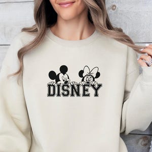 K&ouml;nnte beinhalten: Sandfarbenes Sweatshirt mit einer schwarz-wei&szlig;en Grafik von Mickey und Minnie Mouse &uuml;ber dem Wort "DISNEY" in einer College-Schriftart. Das Sweatshirt hat eine neutrale Farbe und einen Rundhalsausschnitt.