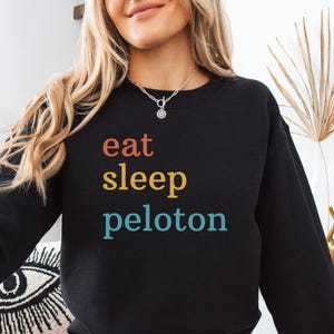 May include: Black crew neck sweatshirt with the words "eat sleep peloton" in orange, yellow, and teal lettering. The sweatshirt is worn by a person wearing denim shorts. A black circle with the word "Black" is in the lower right corner.