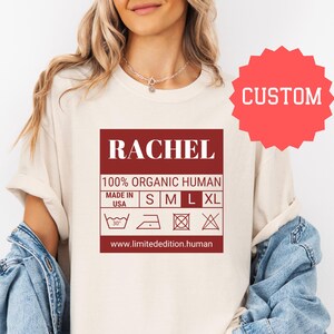May include: A white t-shirt with a red label that says "RACHEL 100% ORGANIC HUMAN MADE IN USA S M L XL www.limitededition.human". The label has a graphic of a washing instruction tag with symbols for washing, ironing, and drying.