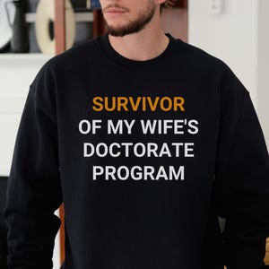 Könnte beinhalten: Schwarzer Pullover mit weißem Text, der "SURVIVOR OF MY WIFE'S DOCTORATE PROGRAM" lautet. Der Text ist orange.