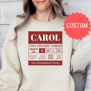 May include: A beige sweatshirt with a red label that says "CAROL 100% ORGANIC HUMAN MADE IN USA S M L XL" with a care label underneath. The label also includes a website address: www.limitededition.human. The sweatshirt is on a model wearing blue jeans.