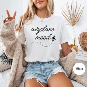 以下が含まれることがあります： 「airplane mood」というテキストと小さな飛行機のグラフィックが描かれた白いTシャツ。長いブロンドの髪をした人が着ています。