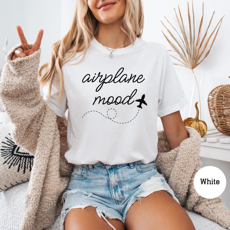 以下が含まれることがあります： 「airplane mood」というテキストと小さな飛行機のグラフィックが描かれた白いTシャツ。長いブロンドの髪をした人が着ています。