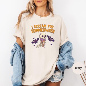 May include: Ivory-colored t-shirt with the text "I Scream for Summerween" in orange. The graphic features a skull holding an ice cream cone, with bats and purple accents. A casual, Halloween-themed tee.