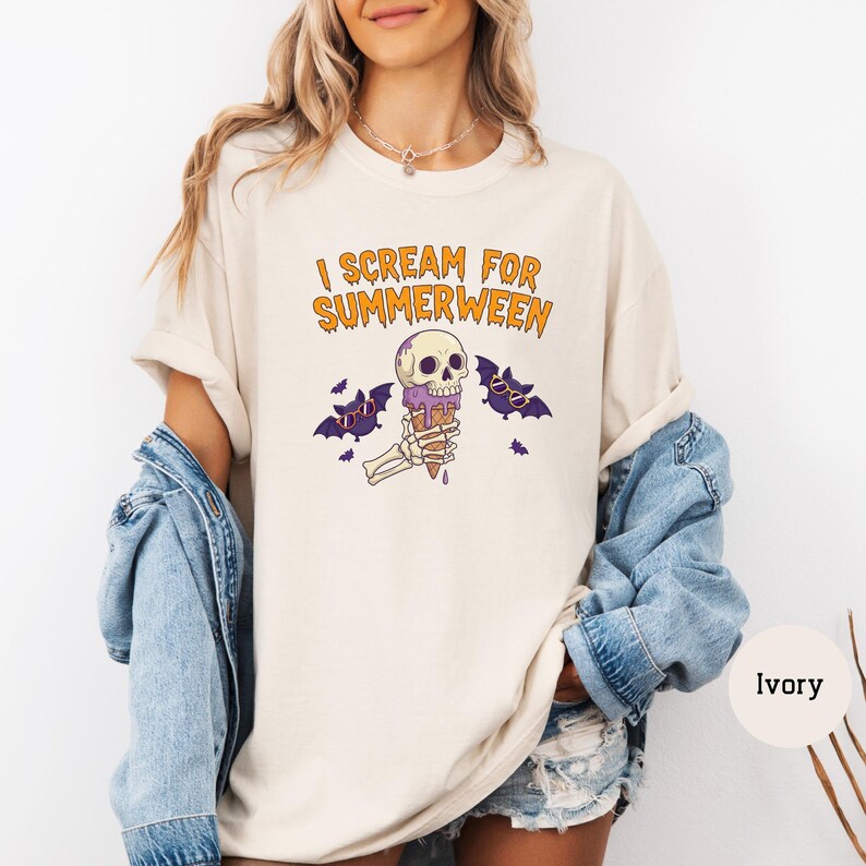 May include: Ivory-colored t-shirt with the text "I Scream for Summerween" in orange. The graphic features a skull holding an ice cream cone, with bats and purple accents. A casual, Halloween-themed tee.