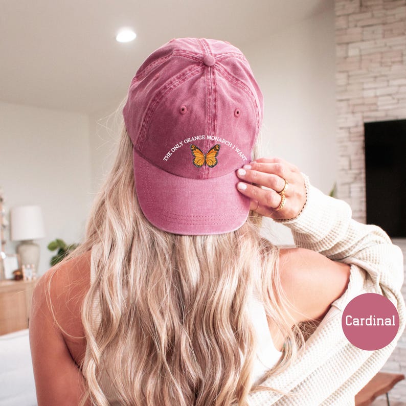 May include: A cardinal red baseball cap with the text "THE ONLY ORANGE MONARCH I WANT" and a butterfly graphic. The cap is made of a soft, textured material and has a curved brim. The cap is a stylish accessory.