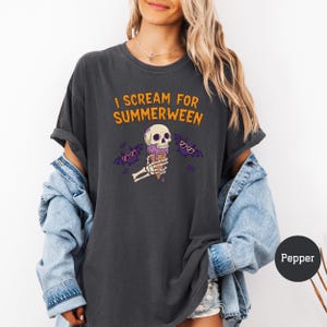 May include: Dark gray t-shirt with the text "I Scream for Summerween" in orange. The graphic features a skull holding an ice cream cone, with purple bats wearing sunglasses. The shirt is a casual, oversized fit.