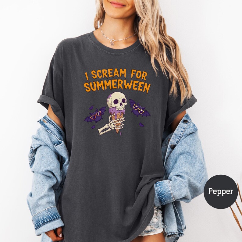 May include: Dark gray t-shirt with the text "I Scream for Summerween" in orange. The graphic features a skull holding an ice cream cone, with purple bats wearing sunglasses. The shirt is a casual, oversized fit.