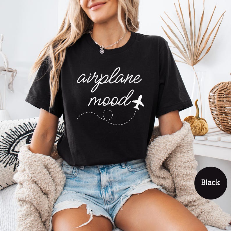以下が含まれることがあります： 白い文字で「airplane mood」と小さな飛行機のグラフィックが書かれた黒いTシャツ。白いカーディガンと青いデニムのショートパンツを着用した人が着ています。