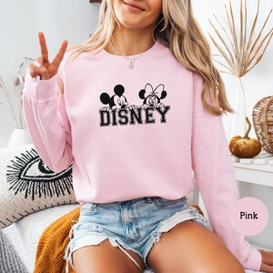 K&ouml;nnte beinhalten: Ein hellrosa Sweatshirt mit einer schwarz-wei&szlig;en Grafik von Mickey und Minnie Mouse &uuml;ber dem Wort "DISNEY". Das Sweatshirt hat lange &Auml;rmel und einen Rundhalsausschnitt. Das Wort "Pink" befindet sich in einem rosa Kreis unten rechts.