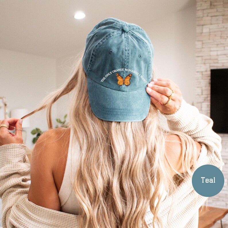May include: Teal baseball cap with the text "THE ONLY ORANGE MONARCH I WANT" and a butterfly graphic. The cap is made of a soft material and has a curved brim. A casual accessory for everyday wear.