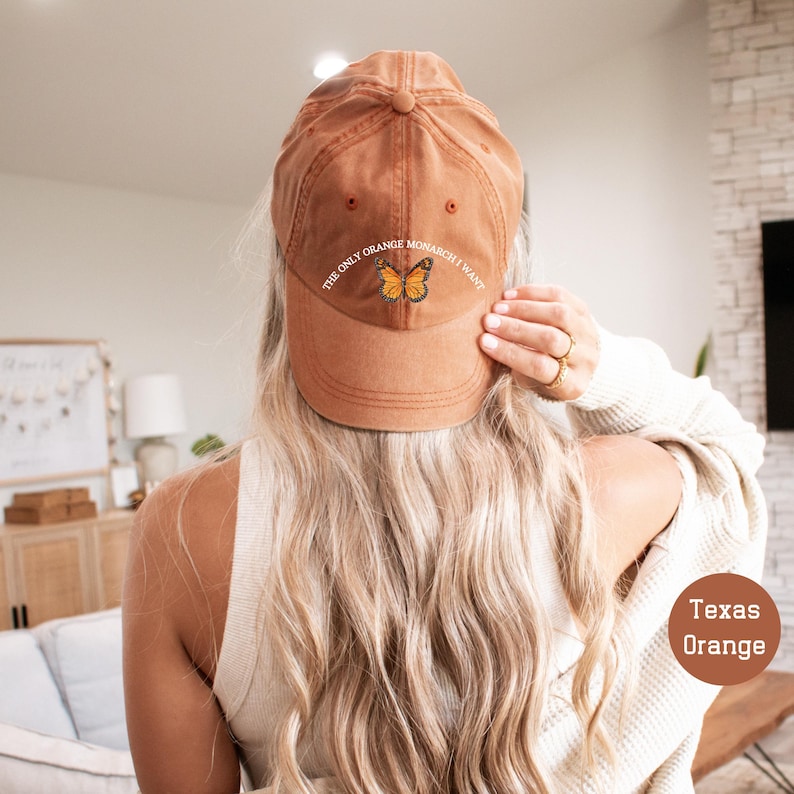 May include: A Texas Orange baseball cap with the text "THE ONLY ORANGE MONARCH I WANT" and a butterfly graphic. The cap is a solid orange color and has a curved brim. The cap is made of a soft material.