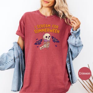 May include: Crimson-colored t-shirt with the text "I Scream for Summerween" in orange. The graphic features a skull-shaped ice cream cone held by a skeleton hand, with purple bats wearing sunglasses. A denim jacket is draped over the arm.