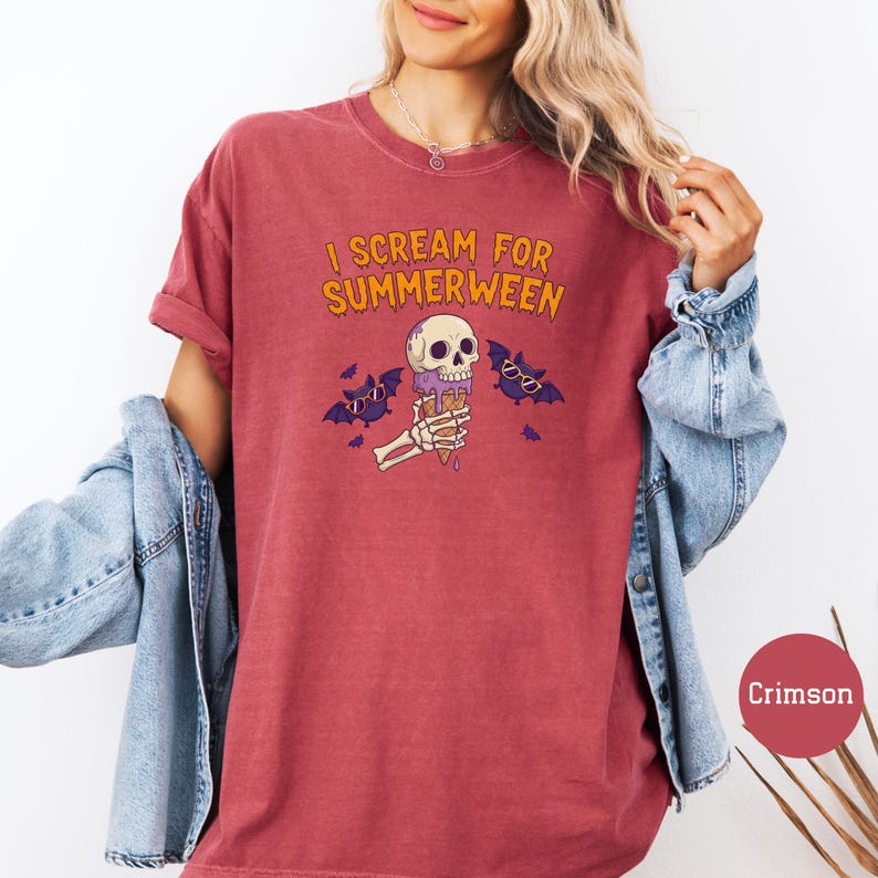 May include: Crimson-colored t-shirt with the text "I Scream for Summerween" in orange. The graphic features a skull-shaped ice cream cone held by a skeleton hand, with purple bats wearing sunglasses. A denim jacket is draped over the arm.