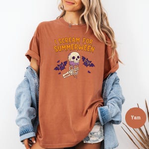 May include: A rust-colored t-shirt with the text "I Scream for Summerween" in orange. The graphic features a skull ice cream cone with purple dripping ice cream, skeleton hands, and purple bats. The shirt is a comfortable, casual style.