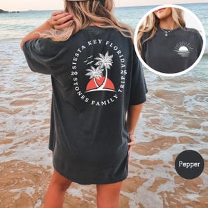 Może przedstawiać: Czarny, luźny t-shirt z białym nadrukiem na plecach. Nadruk przedstawia okrąg z tekstem "Siesta Key Florida Family Trip 20 Stones 25 Trip" i palmę z czerwonym zachodem słońca za nią. Koszulkę nosi osoba stojąca na plaży.