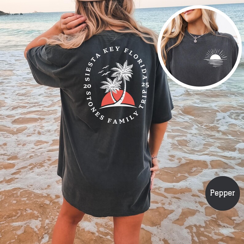 Może przedstawiać: Czarny, luźny t-shirt z białym nadrukiem na plecach. Nadruk przedstawia okrąg z tekstem "Siesta Key Florida Family Trip 20 Stones 25 Trip" i palmę z czerwonym zachodem słońca za nią. Koszulkę nosi osoba stojąca na plaży.