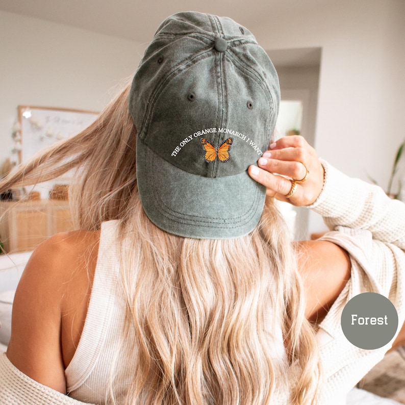 May include: A forest green baseball cap with the text "THE ONLY ORANGE MONARCH I WANT" and an embroidered orange butterfly. The cap is made of a soft, worn material.