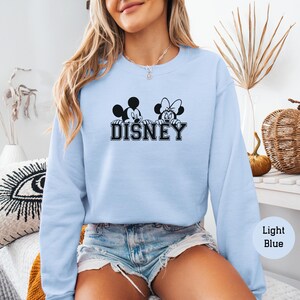 K&ouml;nnte beinhalten: Hellblaues Sweatshirt mit einem schwarzen und wei&szlig;en Motiv von Mickey und Minnie Mouse &uuml;ber dem Wort "DISNEY". Das Sweatshirt hat einen Rundhalsausschnitt und lange &Auml;rmel. Das Bild zeigt auch Jeansshorts. Der Text "Light Blue" befindet sich in einem Kreis.