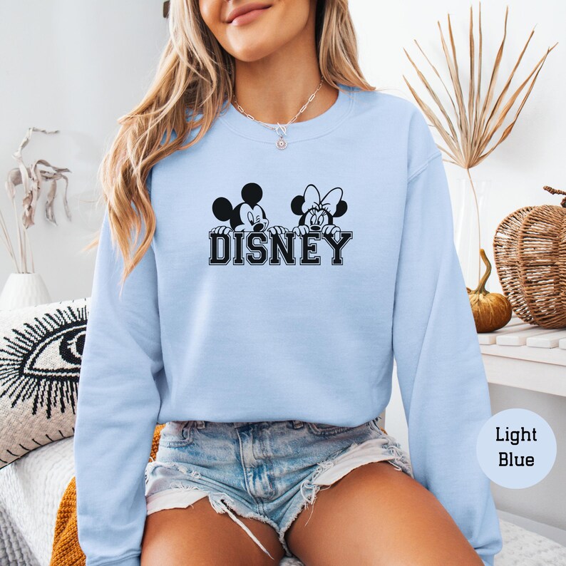 K&ouml;nnte beinhalten: Hellblaues Sweatshirt mit einem schwarzen und wei&szlig;en Motiv von Mickey und Minnie Mouse &uuml;ber dem Wort "DISNEY". Das Sweatshirt hat einen Rundhalsausschnitt und lange &Auml;rmel. Das Bild zeigt auch Jeansshorts. Der Text "Light Blue" befindet sich in einem Kreis.
