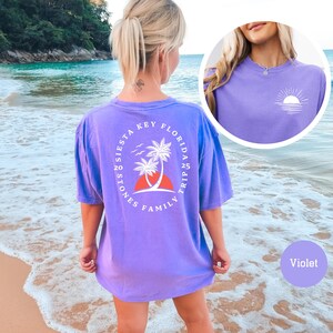 Może przedstawiać: Fioletowy T-shirt z białym nadrukiem z tyłu, kt&oacute;ry głosi "Family Trip 25 Florida Key Siesta Stones". Nadruk przedstawia palmy i zach&oacute;d słońca. Koszulka jest pokazana na modelce stojącej na plaży.