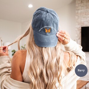 May include: Navy blue baseball cap with the text "THE ONLY ORANGE MONARCH I WANT" and a small embroidered orange butterfly. The cap is made of a soft, worn material.