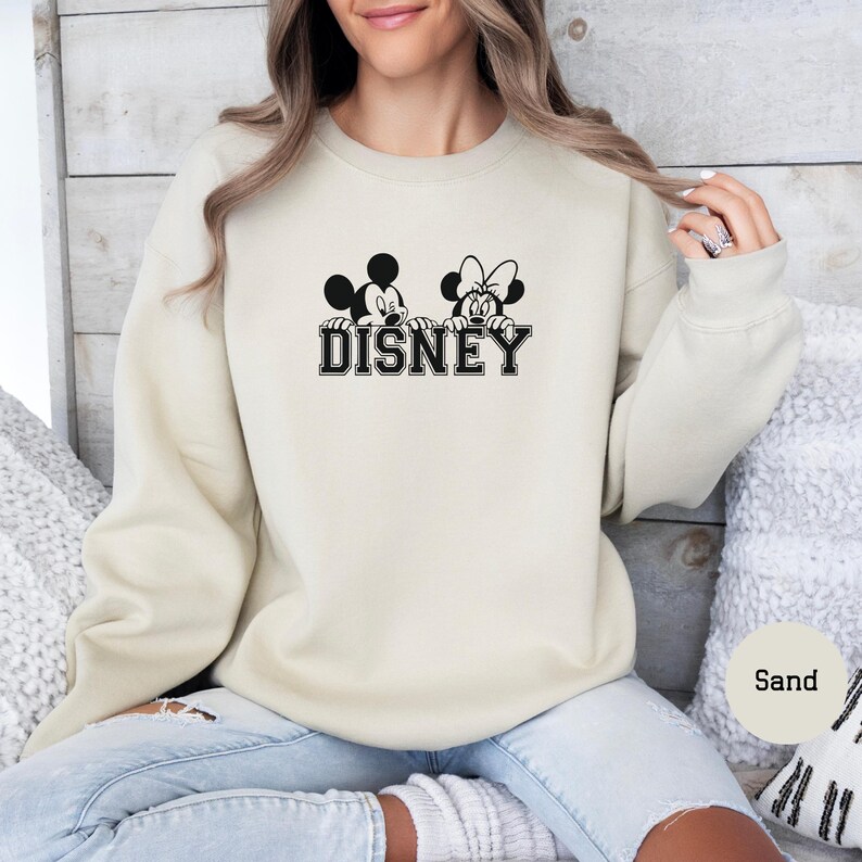 K&ouml;nnte beinhalten: Sandfarbenes Sweatshirt mit einer schwarz-wei&szlig;en Grafik von Mickey und Minnie Mouse &uuml;ber dem Wort "DISNEY" in einer College-Schriftart. Das Sweatshirt hat eine neutrale Farbe und einen Rundhalsausschnitt.