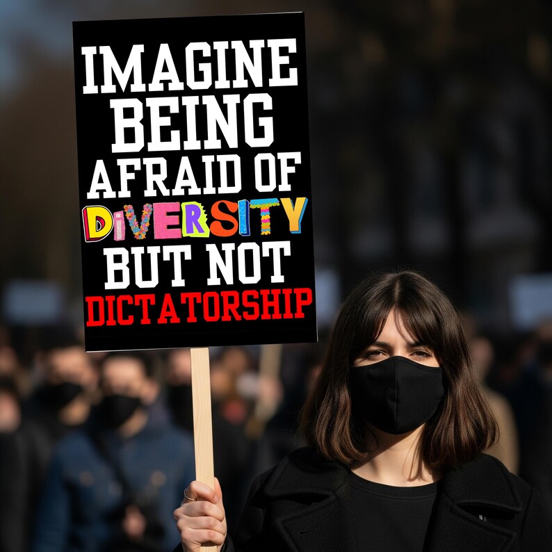 May include: A black sign with the text "IMAGINE BEING AFRAID OF DIVERSITY BUT NOT DICTATORSHIP" in white, red, and colorful letters. A person wearing a black face mask holds the sign in a crowd.