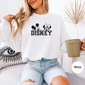 K&ouml;nnte beinhalten: Wei&szlig;es Sweatshirt mit langen &Auml;rmeln und einer schwarz-wei&szlig;en Grafik von Mickey und Minnie Mouse sowie dem Wort "DISNEY". Das Sweatshirt hat einen Rundhalsausschnitt. Eine Kaffeetasse mit dem Wort "COFFEE" wird in der Hand gehalten.