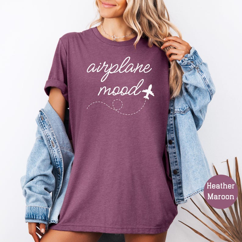 以下が含まれることがあります： ヘザーマロンのTシャツに、「airplane mood」という白い筆記体で書かれた文字。文字の横に白い飛行機のアイコンがあり、その後ろには点線の軌跡がある。