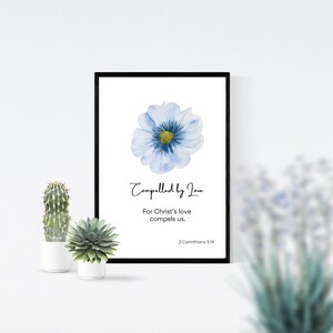 Pode incluir: Uma impressão emoldurada com uma pintura em aquarela de uma flor azul. O texto "Compelled by Love" está escrito em itálico acima da flor. Abaixo da flor, o texto diz "For Christ's love compels us. 2 Corinthians 5:14"