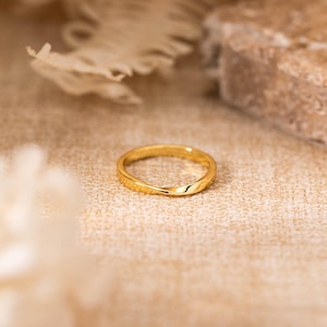 May include: A gold-colored ring with a textured surface. The ring is set against a neutral background with dried flowers and a stone. The ring is a simple band design, suitable for everyday wear.