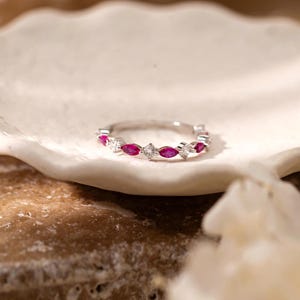 May include: A silver-toned ring featuring an alternating pattern of marquise-cut deep pink stones and small round clear stones set along the band. The metal band has a polished finish, and the stones are arranged in a symmetrical, repeating sequence.