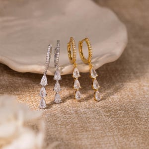 Könnte beinhalten: Zwei Paar Ohrhänger, ein silberfarbenes und ein goldfarbenes. Jeder Ohrring besteht aus einem kleinen, mit winzigen klaren Steinen besetzten Creolen-Ring, an dem drei klare, tropfenförmige Steine in einer vertikalen Linie herabhängen.