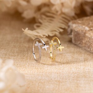 May include: Two open-style rings, one silver-toned and one gold-toned, each featuring a small cross at one end and a band encrusted with tiny clear stones. The rings are positioned upright, showcasing their delicate, adjustable, and minimalist design.