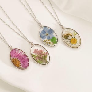 May include: Four silver-toned necklaces with oval pendants, each containing a different dried flower. The flowers are pink, light pink, blue, and white with yellow centers. The pendants are displayed on a white surface.