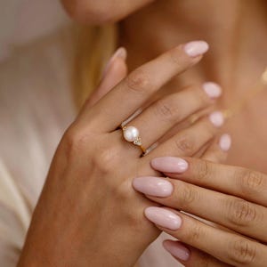 Può includere: Un anello in oro con una perla bianca rotonda centrale, affiancata da due piccole pietre chiare su ciascun lato. La fascia è sottile e metallica, con una finitura lucida. Il design è incentrato sulla perla, fissata saldamente in una montatura d'oro.