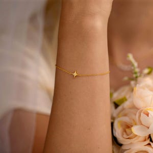 May include: A delicate gold bracelet featuring a starburst charm with a central gemstone. The bracelet is worn on a wrist, showcasing its fine chain and sparkling design. The jewelry is a simple yet elegant accessory.